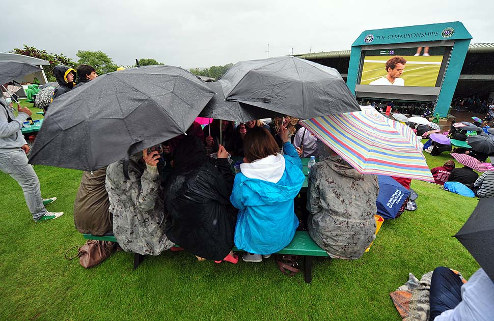 Теннисные фанаты сидят, укрывшись под зонтом, перед большим экраном, по которому идет трансляция с корта