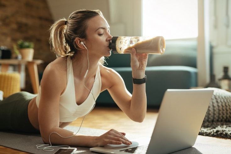 Спортивная девушка пьет протеиновый коктейль и смотрит в ноутбук