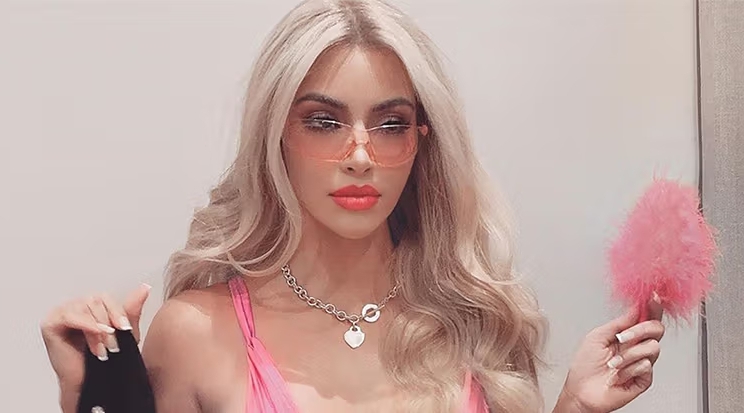 Молодая девушка-бимбо с ярким макияжем, длинными светлыми волосами и розовым аксессуаром в руках