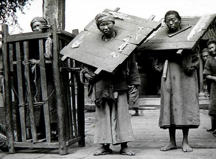 Группа заключённых в тяжёлых деревянных кангах сидит у каменной тюремной стены, Китай, начало XX века.