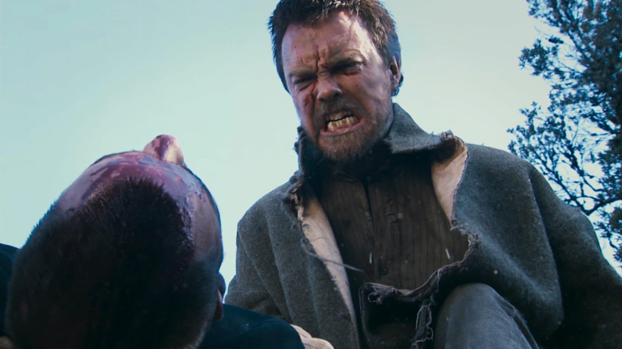 Актер в роли Пирса сидит у костра с диким взглядом, его лицо покрыто грязью и сажей