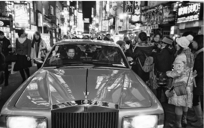 Ч/б фото: Rolls-Royce с улыбающимся мужчиной за рулём стоит в толпе на оживлённой ночной улице Токио, окружён неоновыми вывесками и людьми в зимней одежде