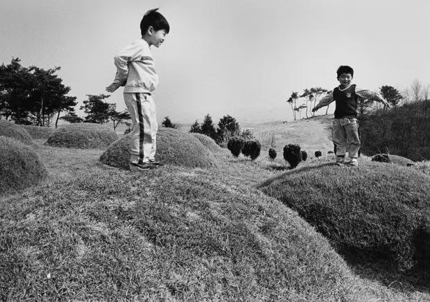 Ч/б фото: двое маленьких мальчиков в спортивной одежде играют на холмистом поле с круглыми подстриженными кустами, прыгают и балансируют на них, на фоне гор и сосен