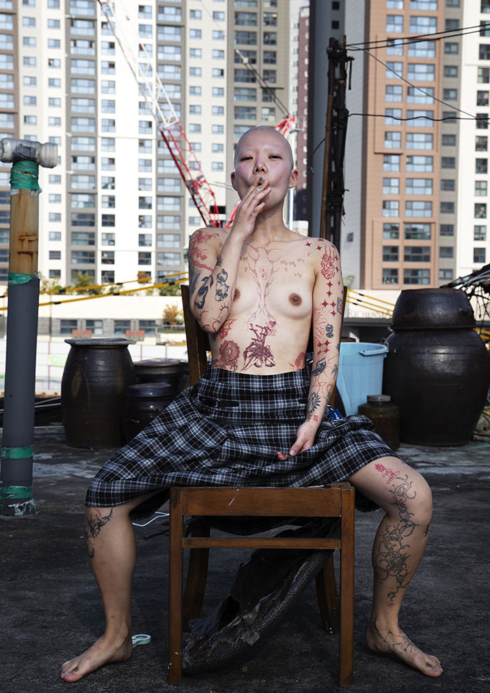 Женщина с бритой головой и обширными красными татуировками по всему телу (включая грудь и ноги) сидит на стуле на крыше, курит сигарету, на фоне высоток и кранов