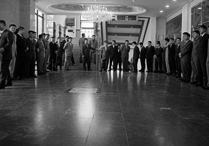 Ч/б фото: группа азиатских мужчин в костюмах стоит в два ряда в роскошном холле с люстрой и мраморным полом, некоторые пожимают руки у центрального постамента