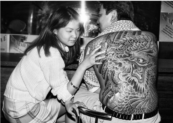 Ч/б фото: молодая азиатская женщина в свитере сидит на корточках в баре, восхищённо трогает рукой огромную татуировку дракона на спине мужчины