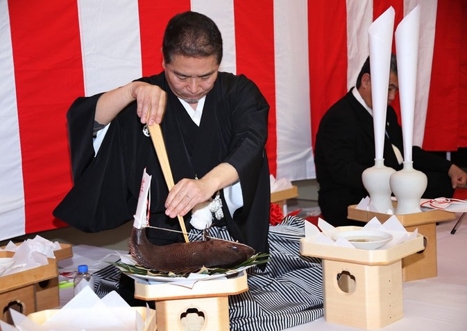 Мужчина в традиционной японской одежде (хаори и хакама) на коленях за низким столом ловко режет большую рыбу деревянными палочками, на фоне красных и белых полос флагов