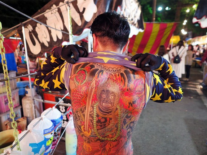 Уличный торговец тэкия с татуировками и в традиционной одежде стоит на фоне ярких вывесок