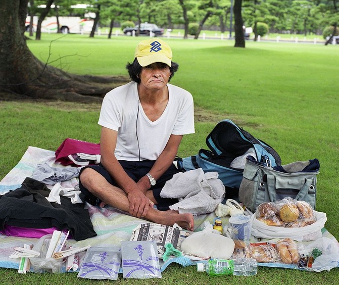 Бездомный мужчина средних лет сидит в парке с сумками и вещами, взгляд в сторону