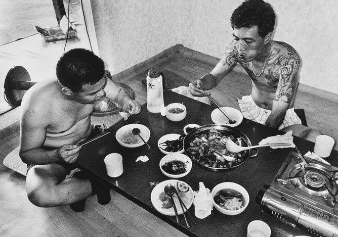 Ч/б фото: два татуированных азиатских мужчины без рубашек едят из общего горячего котла с морепродуктами за низким столом 