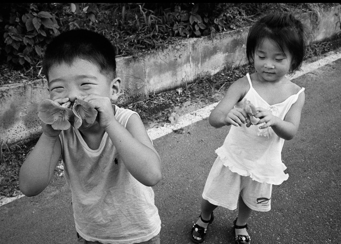 Ч/б фото: двое маленьких азиатских детей на улице &mdash; мальчик в майке нюхает большой цветок, зажмурившись, девочка в платье держит цветок в руках и улыбается