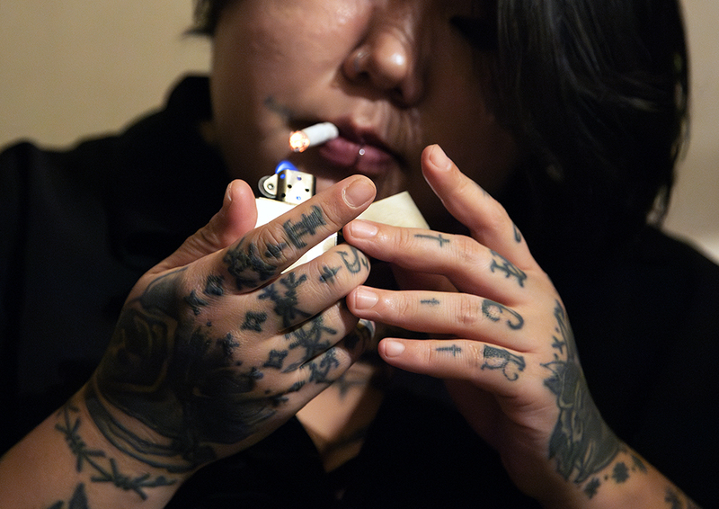 Женщина с татуировками на пальцах, руках и лице прикуривает сигарету зажигалкой, держит её близко к губам с пирсингом, крупный план