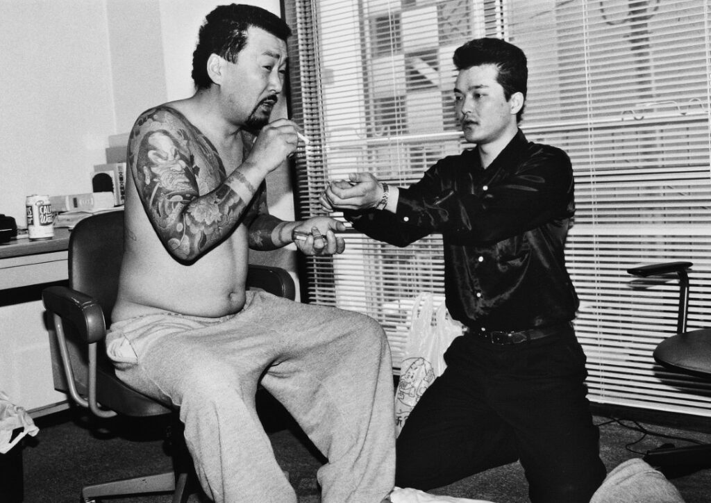 Ч/б фото: якудза с татуировками курит, пока молодой тату-мастер работает над его рукой в офисе с жалюзи