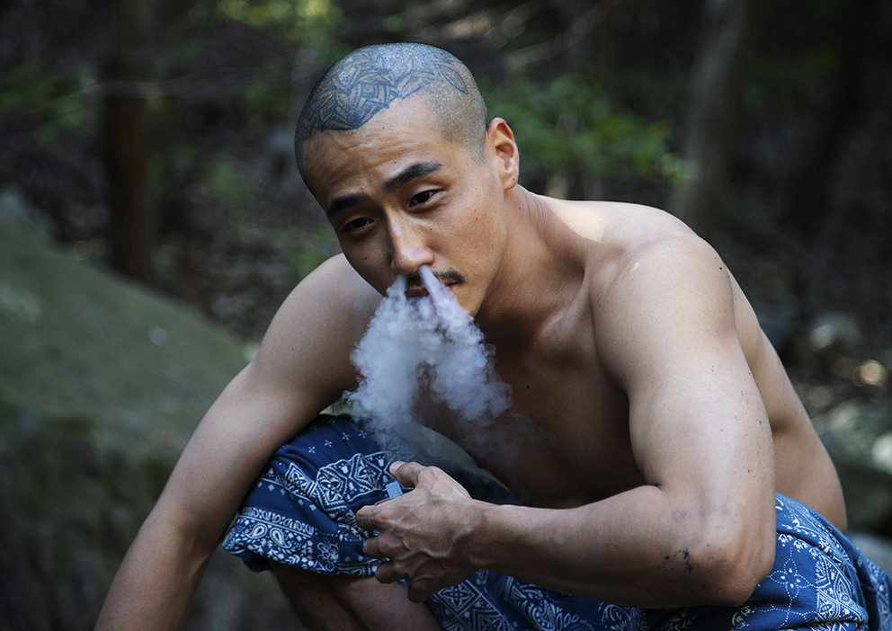 Мускулистый азиатский мужчина с выбритой головой и татуировкой орнамента на затылке курит в лесу, сидя на корточках в синем парео, выдыхает густой дым