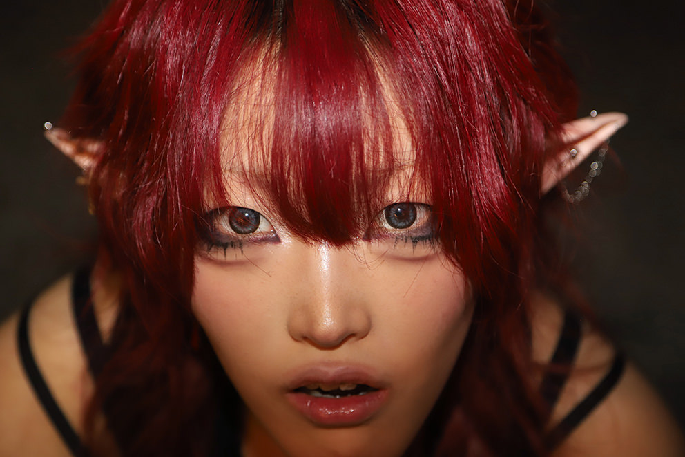 Девушка с ярко-красными волосами, эльфийскими ушами с пирсингом и цепочками, густым макияжем и открытым ртом пристально смотрит в камеру в тёмном помещении