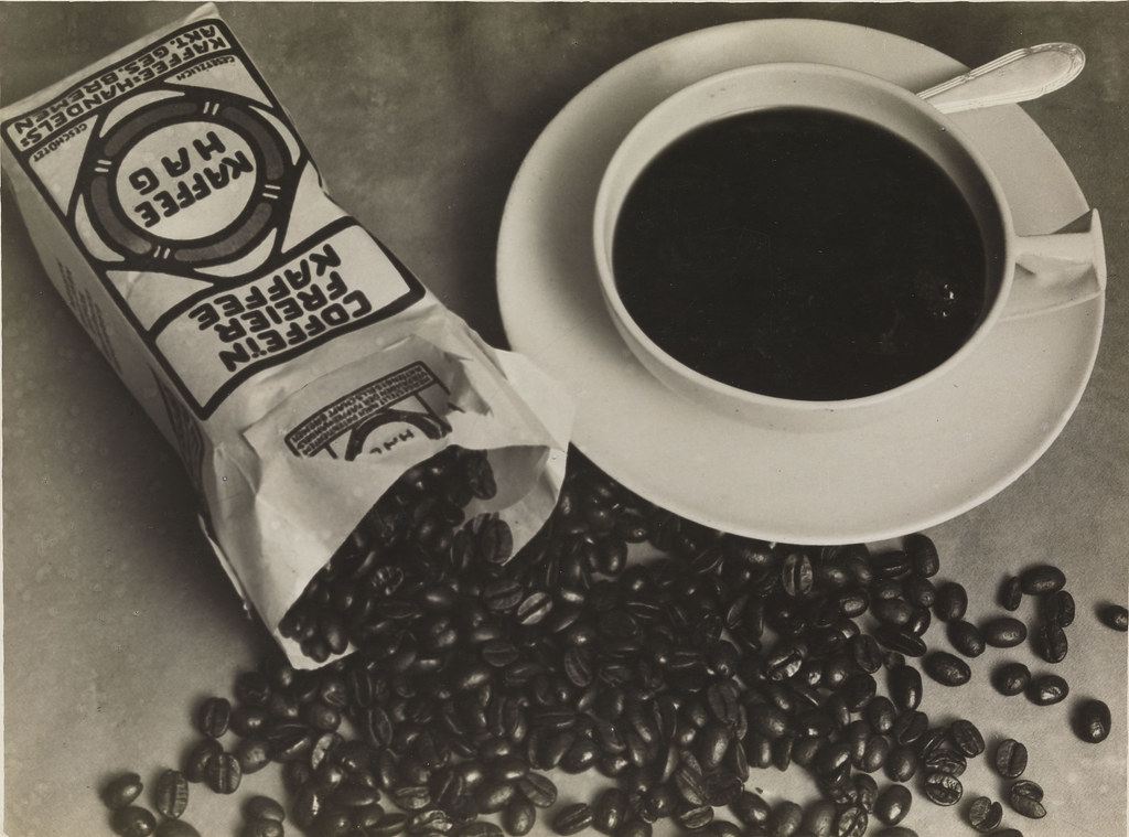 Винтажная рекламная композиция Kaffee HAG: пачка кофе, чашка с дымящимся напитком и горсть кофейных зерен на столе.