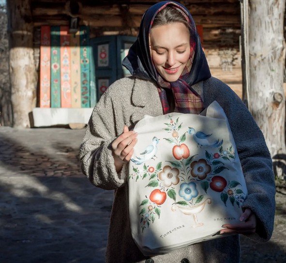 Девушка с расписанной торбой в руках на фоне традиционного деревянного дома