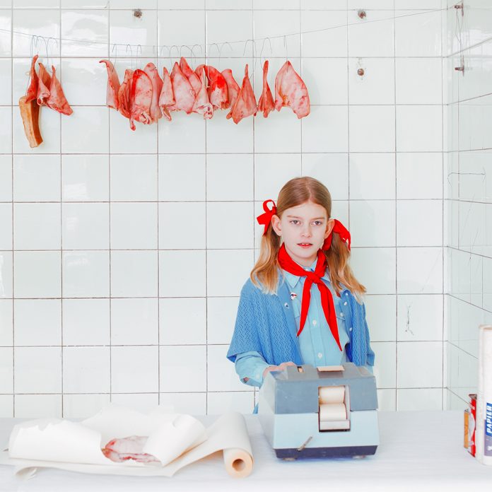 Девочка в синей блузке стоит за мясным прилавком с колбасами и мясом, висящим на крюках
