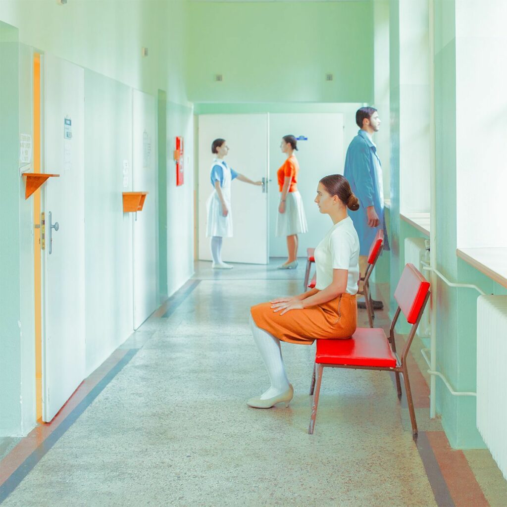 Женщина в оранжевой юбке сидит на красном стуле в коридоре больницы с медсёстрами