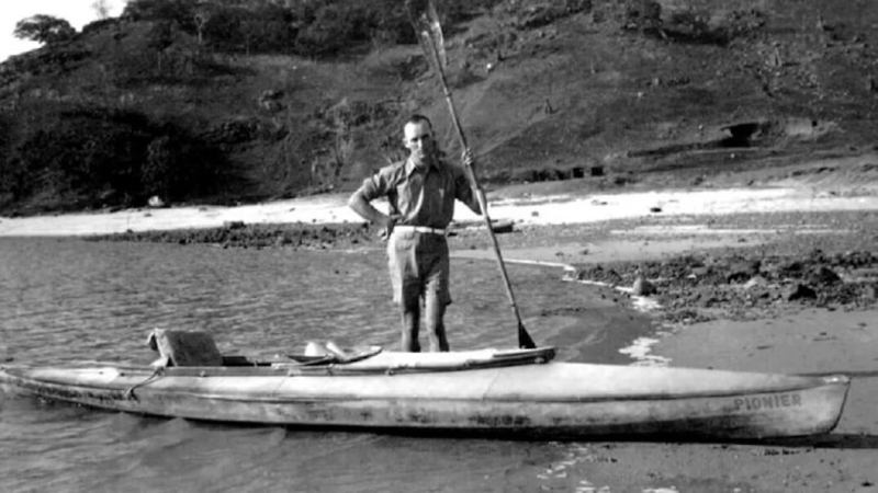 Оскар Спек с веслом стоит возле складной байдарки на песчаном берегу