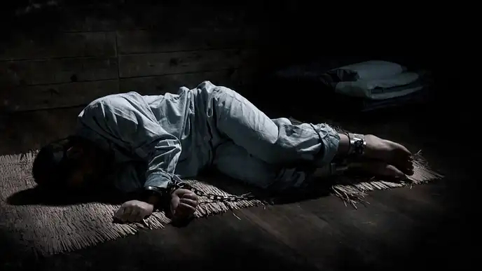 Человек с закованными руками и ногами лежит в тёмном помещении на циновке, реконструкция условий в тюрьме Абасири