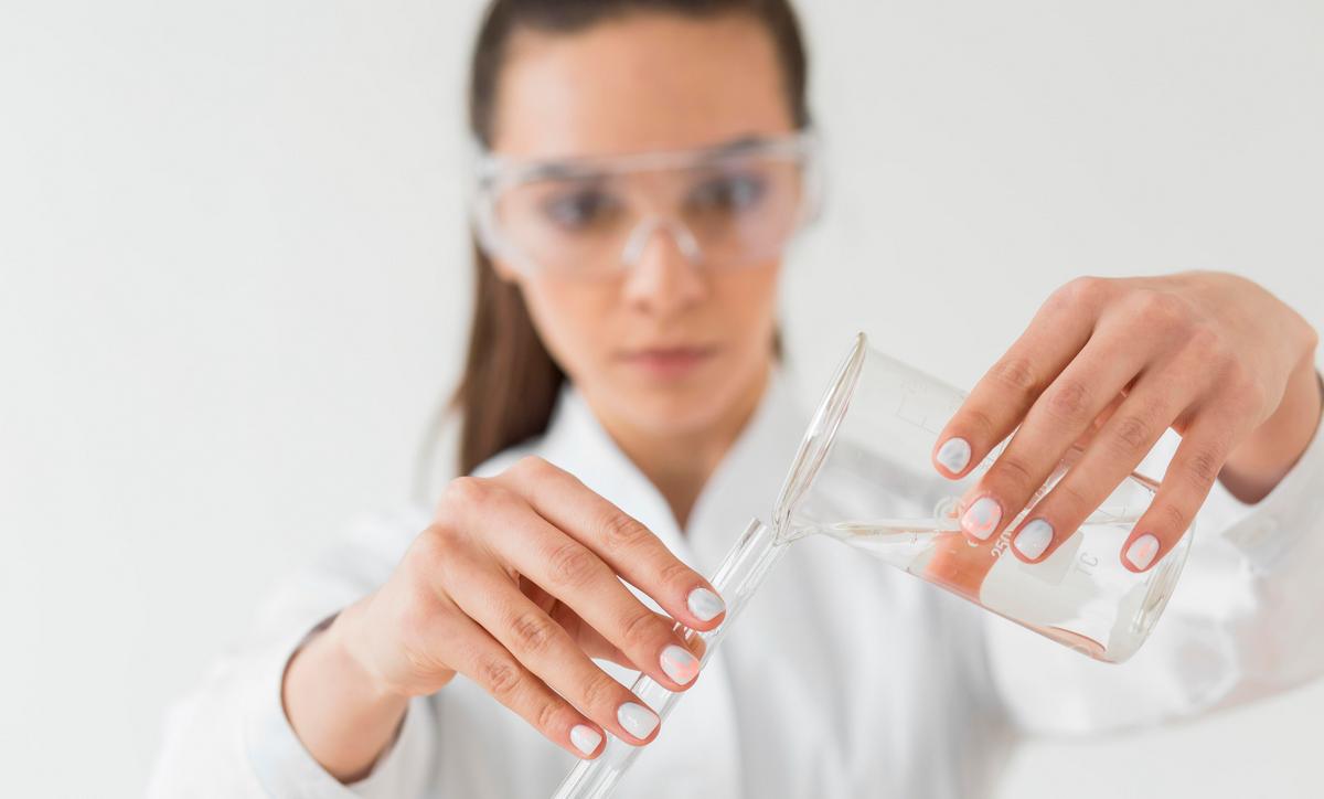 Женщина-лаборант в защитных очках переливает чистую воду из колбы в другой сосуд в научной лаборатории