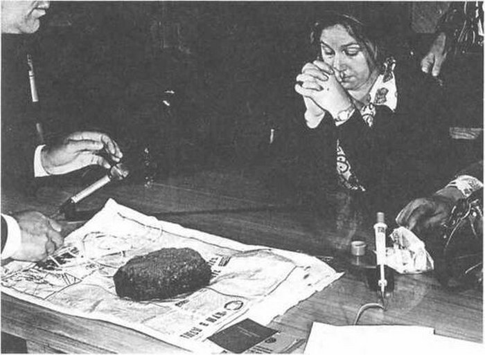 Марта Петерсон сидит за столом, перед ней тайник в виде куска асфальта