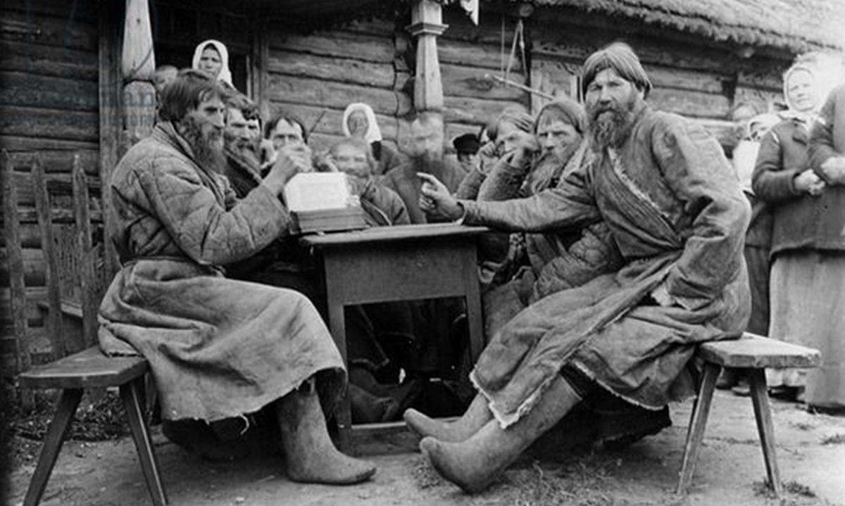 Группа бородатых крестьян в валенках и традиционной одежде сидит за столом у деревянной избы на фоне толпы