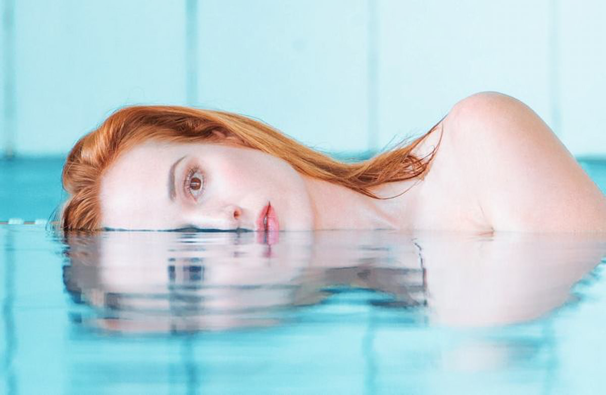 Женщина с рыжими волосами лежит лицом вверх в бассейне с голубой водой, создавая зеркальное отражение