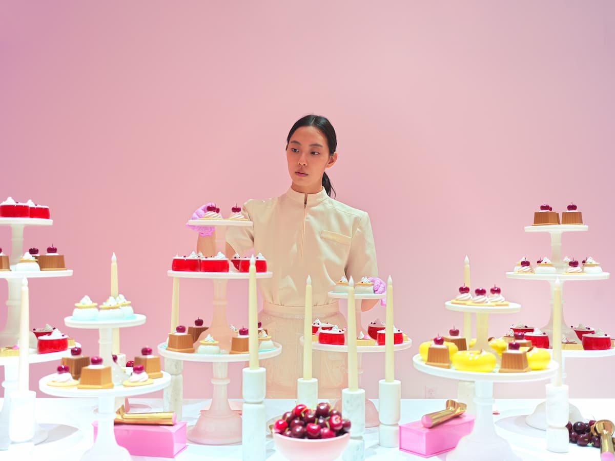 Женщина в бежевом костюме стоит среди десертов и свечей на розовом фоне 