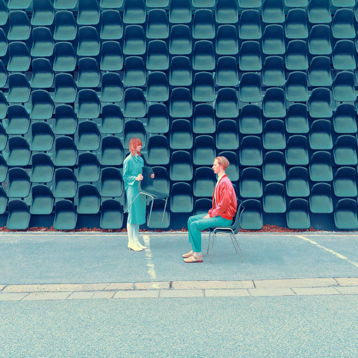 Женщина в бирюзовом платье и мужчина в красной куртке сидят на стульях напротив рядов синих сидений стадиона