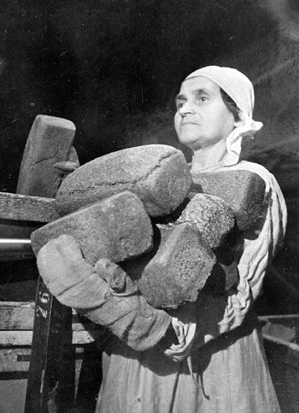 Женщина в платке держит в руках несколько буханок тяжелого блокадного хлеба на фоне печи
