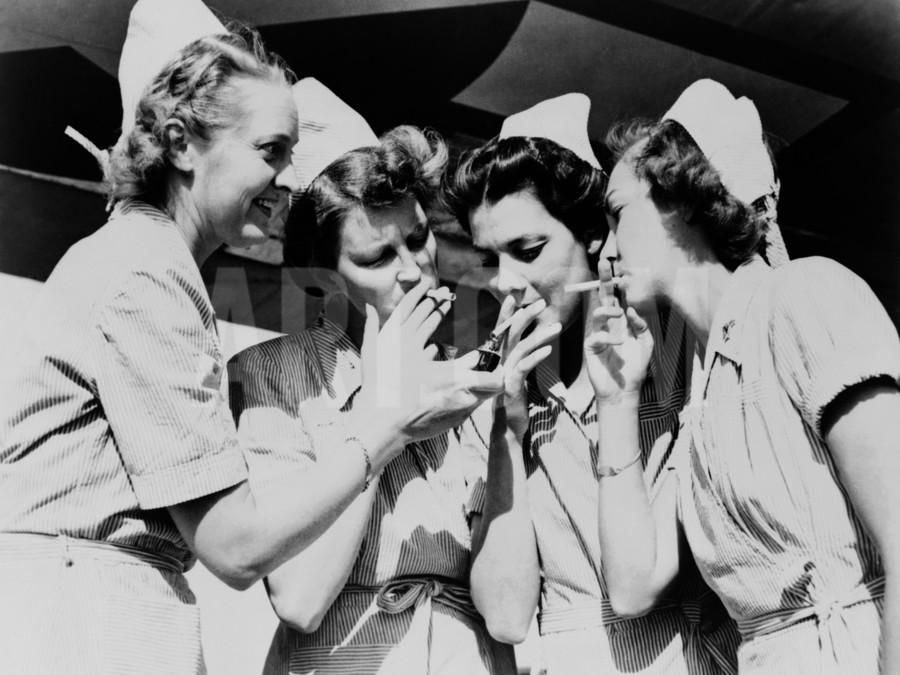 Две медсестры в белых халатах курят, сидя на стульях в больничном коридоре