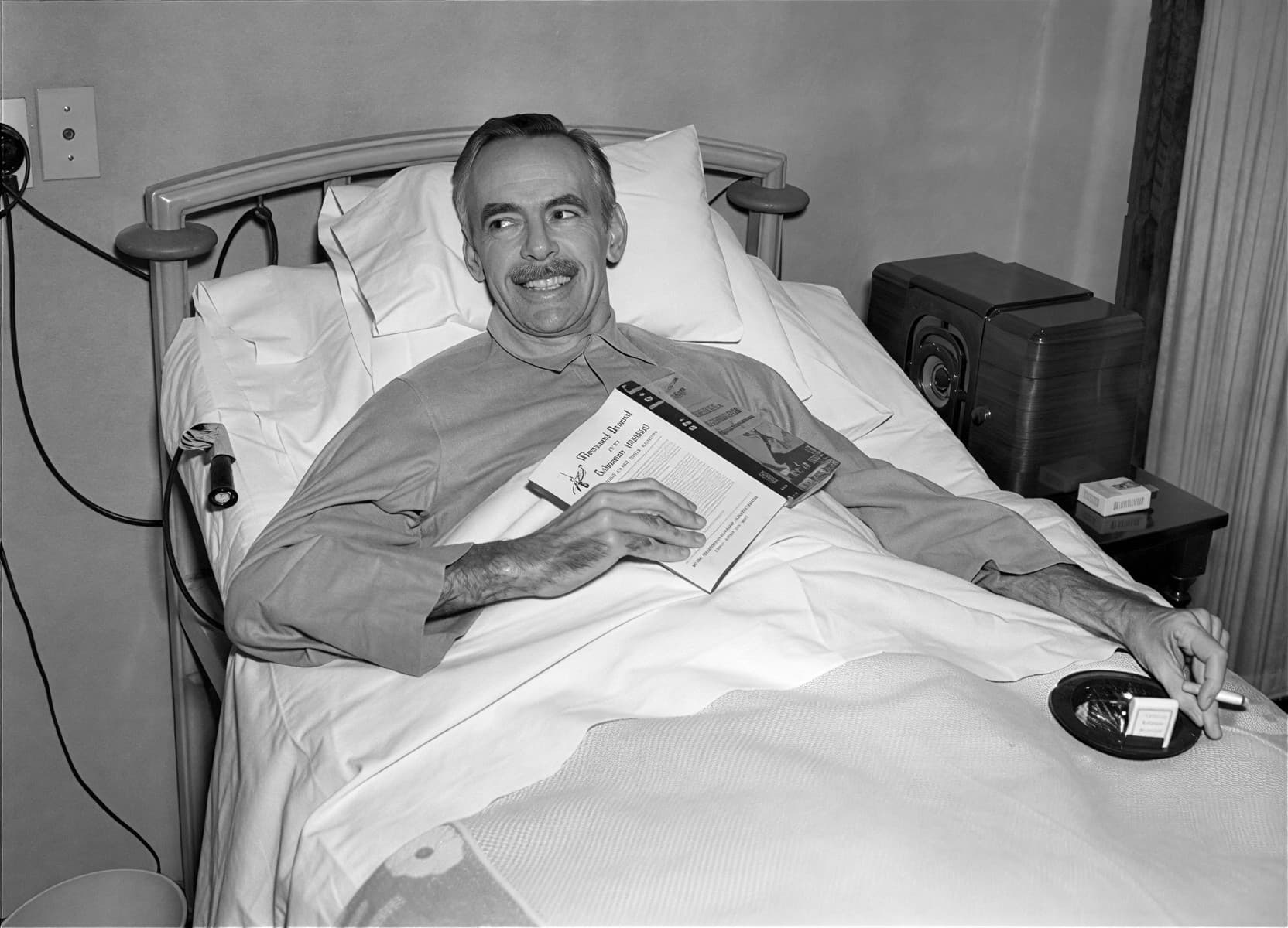 Пожилой пациент в больничной койке читает книгу и курит сигарету