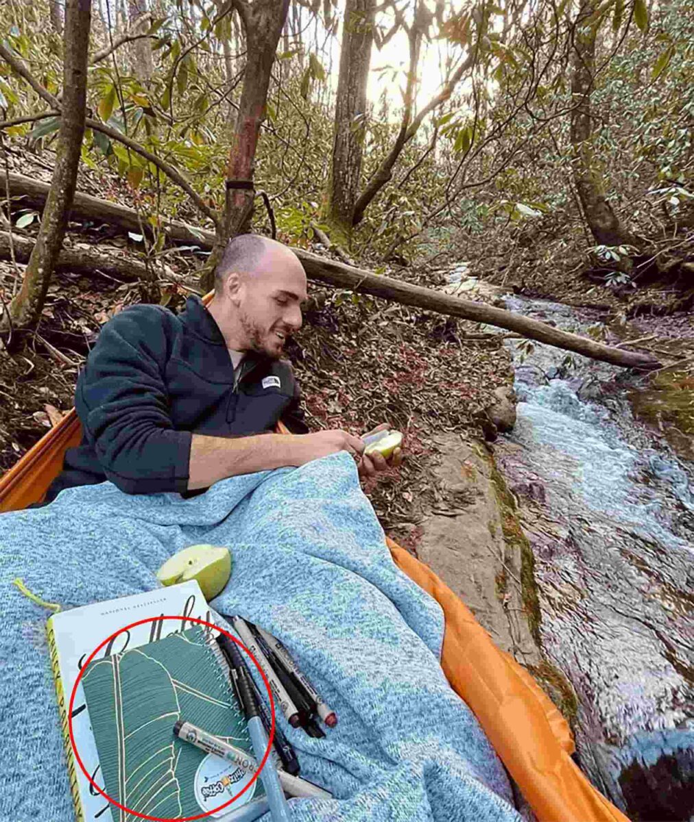 Брайан Лондри сидит в лесу с книгой и маркерами на гамаке у ручья