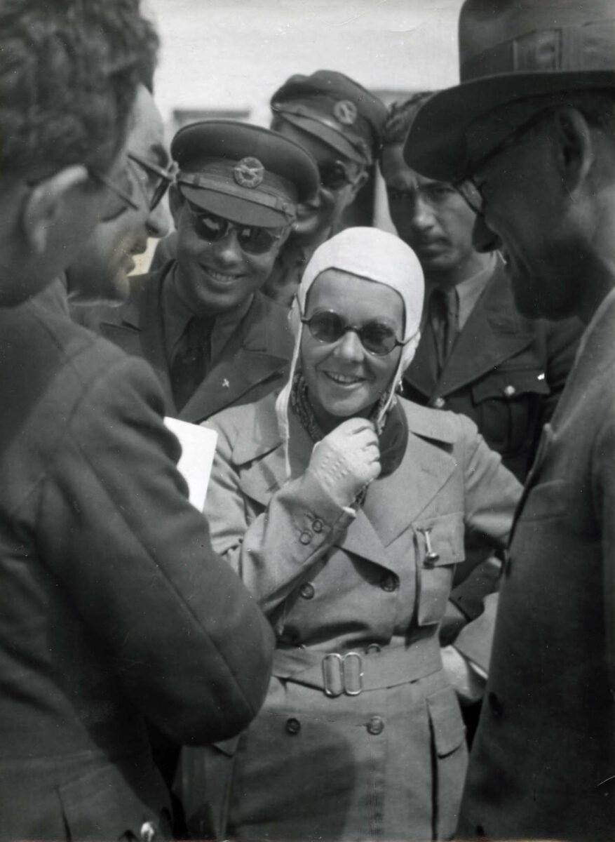 Сабиха Гёкчен в летной форме и шлеме улыбается стоя среди военных