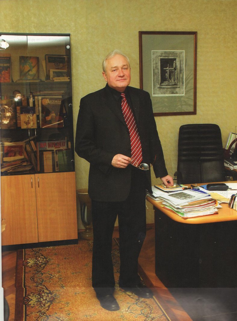 Пожилой мужчина, Николай Гринёв, сидит за рабочим столом в окружении книг