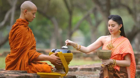 Тайка в традиционной одежде подаёт еду буддийскому монаху