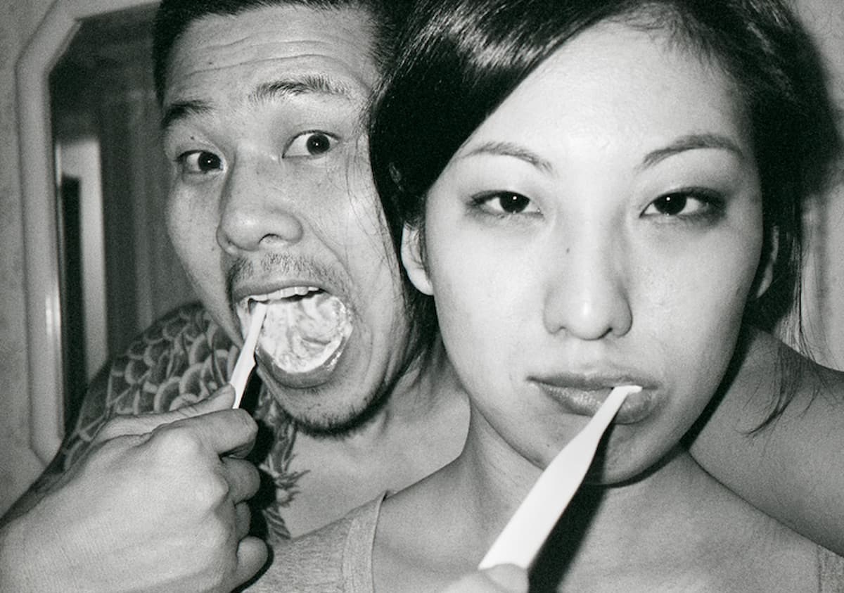 Ч/б фото: азиатская пара в ванной &mdash; мужчина с татуировкой на груди и открытым ртом с зубной щёткой во рту, женщина с макияжем и щёткой во рту