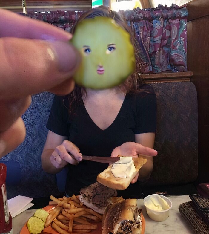 Рука держит огурец с нарисованным лицом перед девушкой за столом