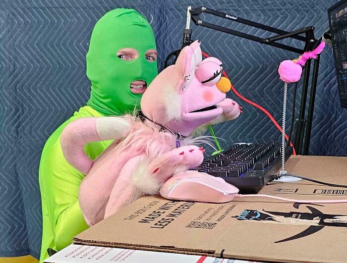 Мужчина в зелёной балаклаве обнимает большую розовую плюшевую кошку за компьютером