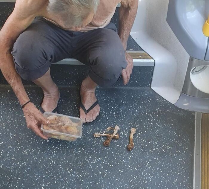 Пожилой мужчина без рубашки сидит на корточках в транспорте с контейнером еды и куриными костями на полу