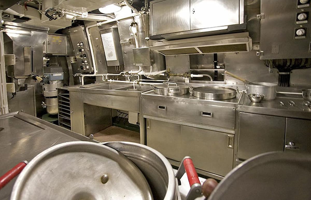 Камбуз подводной лодки: повар в робе разливает суп по мискам у горячей плиты.