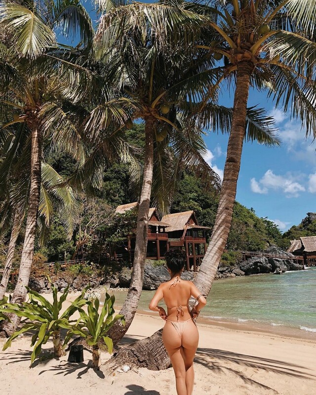 Девушка с упругой попой в бежевом бикини стоит среди пальм на тропическом пляже