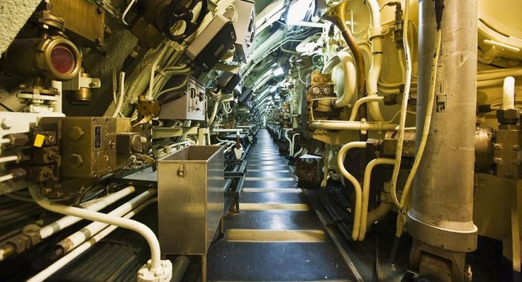 Узкий технический коридор подводной лодки, заставленный оборудованием и трубопроводами. По нему идет моряк.