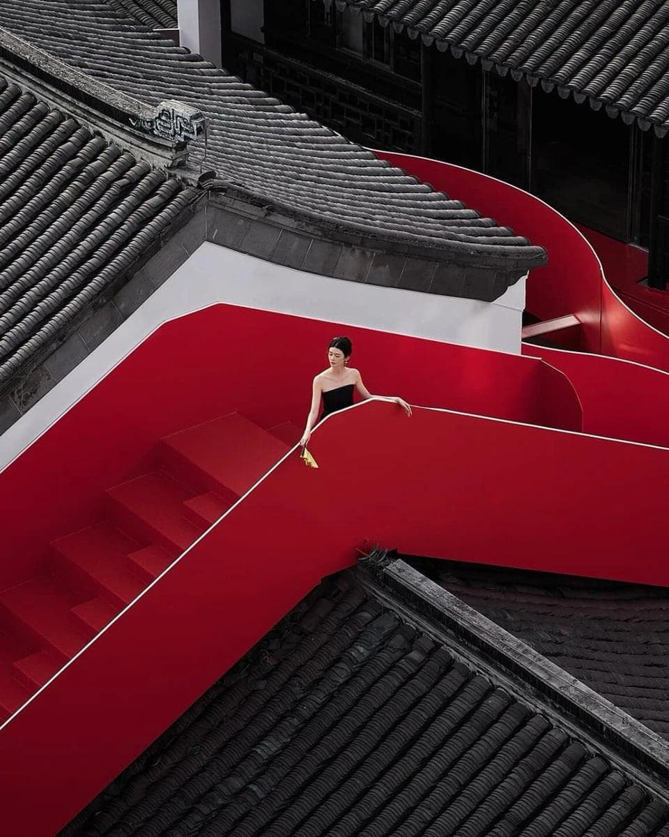Девушка в чёрном платье стоит на красной изогнутой лестнице среди чёрных крыш