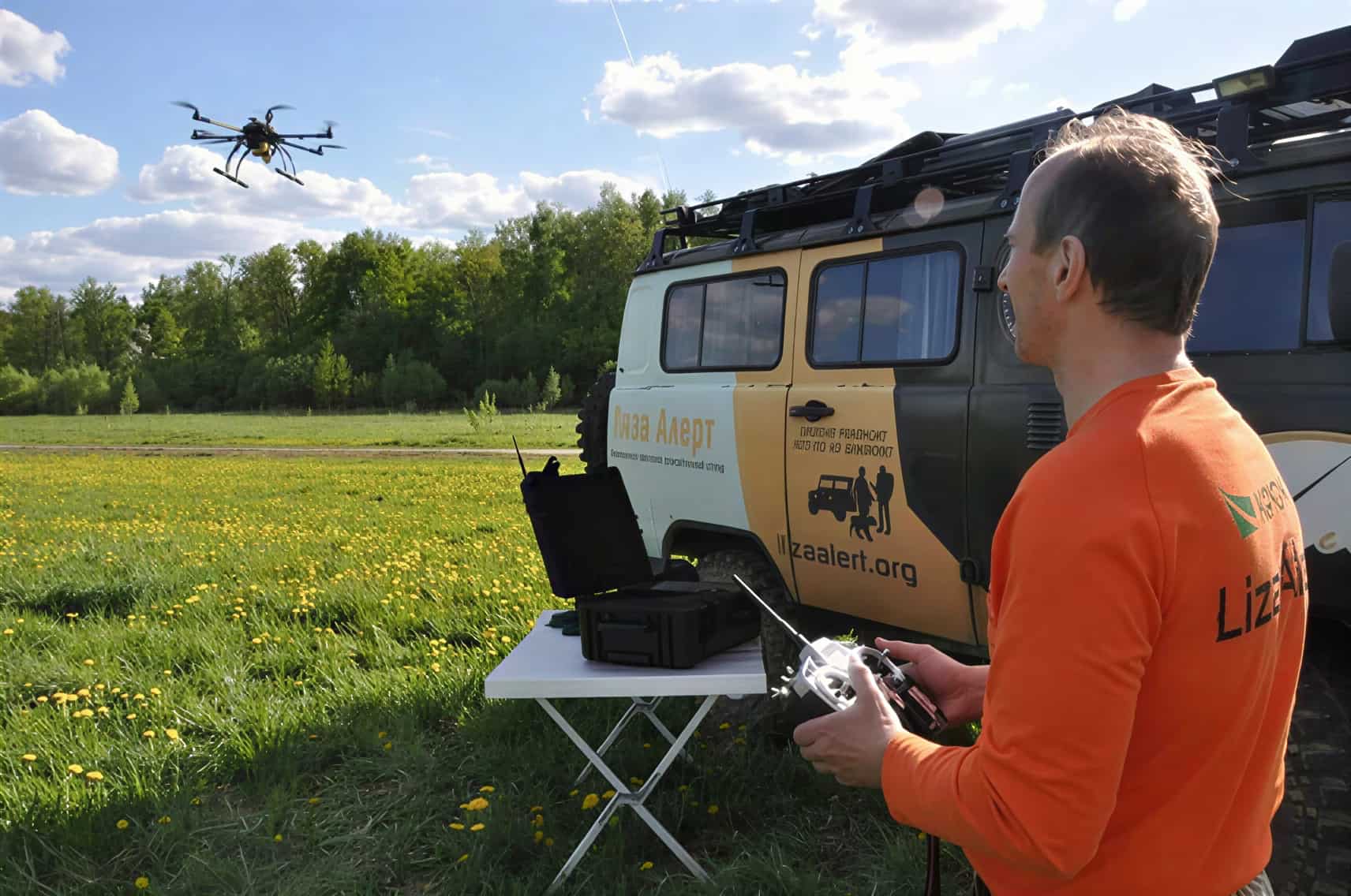 Мужчина в зимней куртке и шапке смотрит в экран планшета, подключённого к пульту дрона, на фоне заснеженных елей