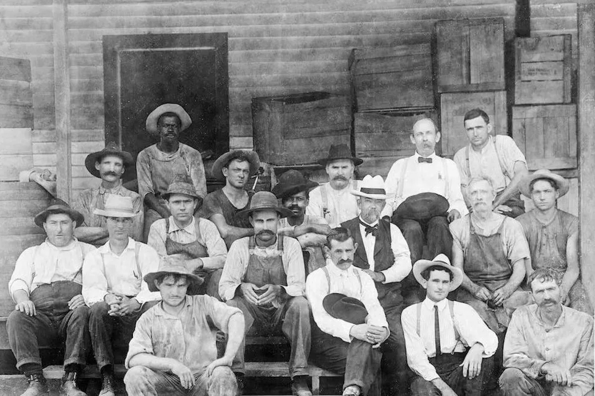 Джек Дэниел в белой шляпе и с усами позирует на фоне дистиллерии вместе с Джорджем Грином, сыном мастера Нейреста Грина