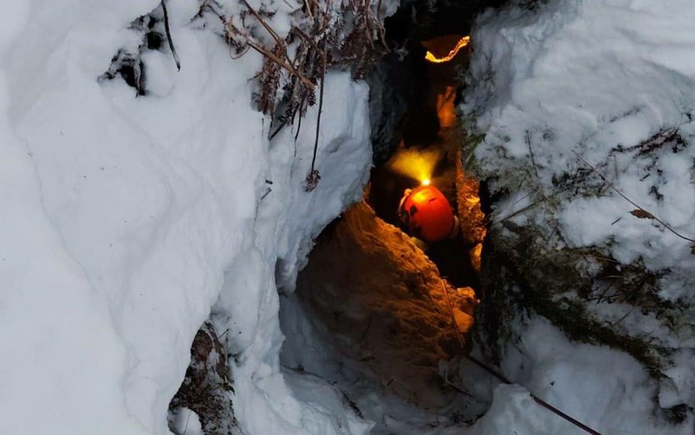 Спелеолог в защитной каске и обвязке спускается на верёвке в тёмный провал пещеры-колодца среди снегов
