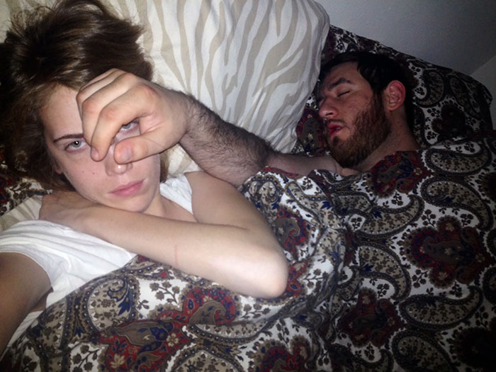 Женщина смотрит в камеру, рядом спит бородатый мужчина в кровати